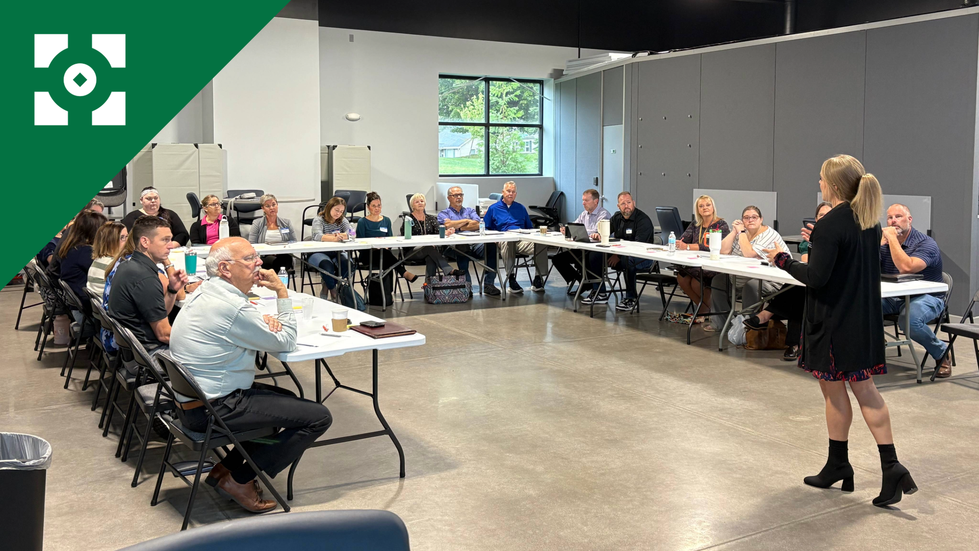 A woman stands and speaks to a seated audience in a conference room with a high ceiling, gray walls, and a large window showing trees outside. Attendees are listening attentively, some taking notes.