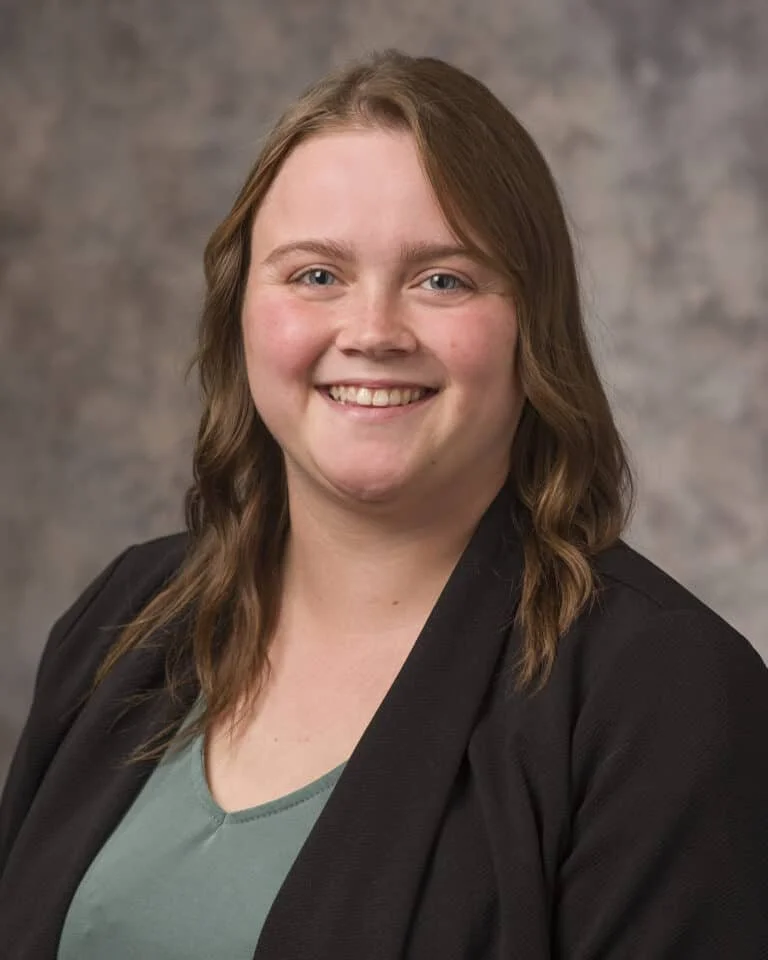 A young woman with shoulder-length brown hair, smiling, wearing a black blazer over a green top, posing against a neutral background.