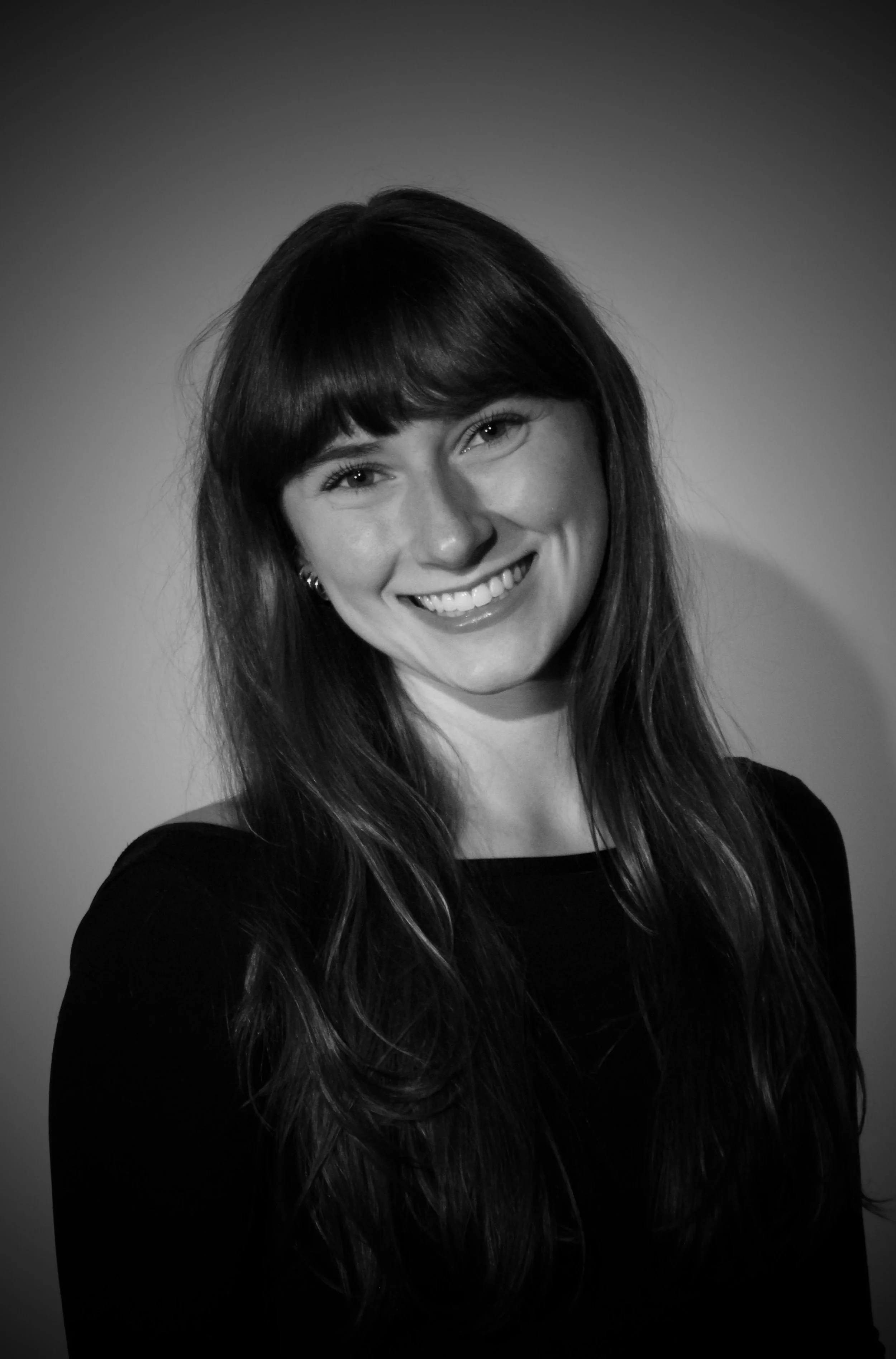 A smiling woman with straight dark hair and bangs, wearing earrings and a black top, in a black and white portrait with a plain background.