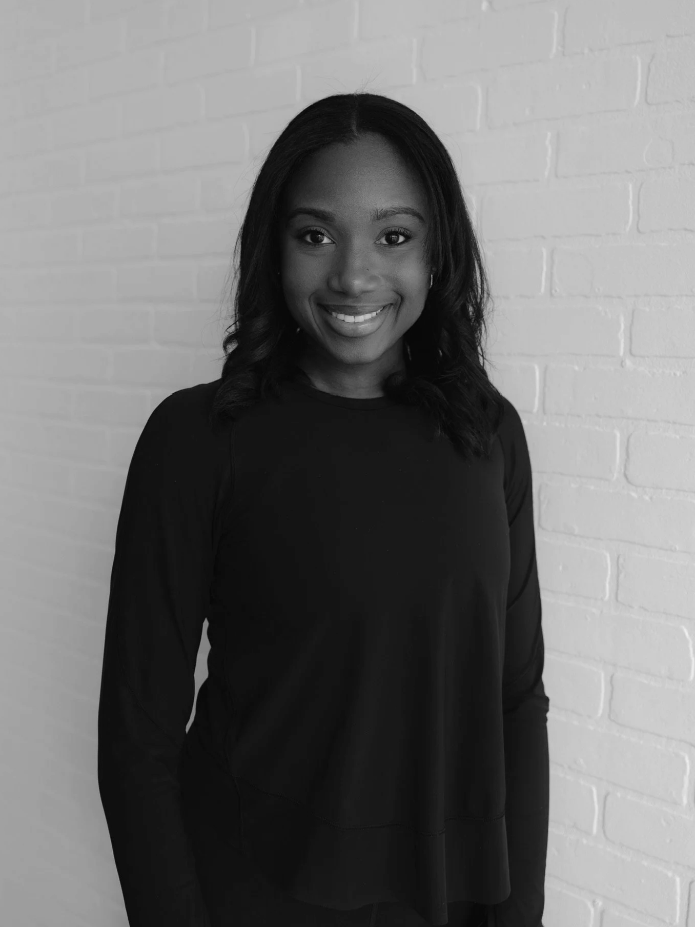 A smiling woman with long black hair, wearing a black long-sleeved shirt, standing against a white brick wall.