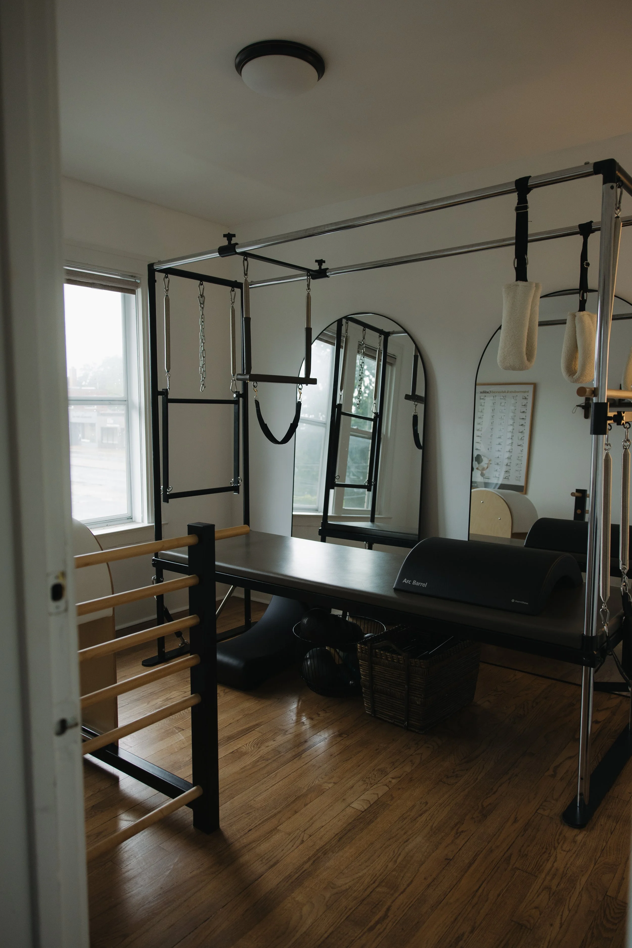 An intimate Pilates room for private instruction with large windows, a mirrored wall, various exercise equipment including a Pilates Cadillac, Barrels and props on the wooden floor. 