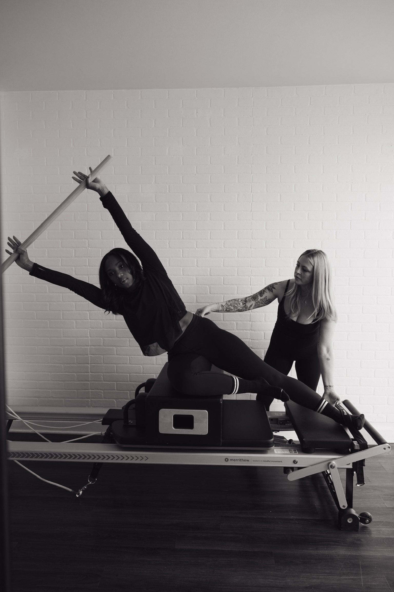 Two women are performing a stretch exercise on a Pilates reformer machine, one lying sideways on the machine with her legs extended and holding a wooden stick, while the other stands beside her assisting. The setting is indoors with a white brick wall in the background.