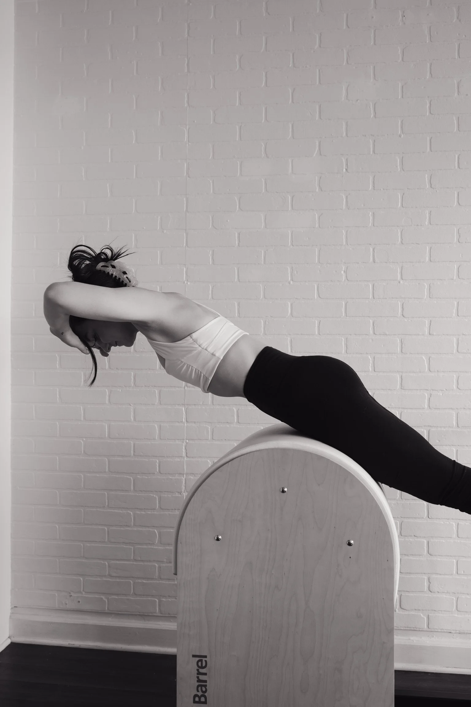 A person is practicing yoga indoors, lying on their stomach on a large cylindrical wooden barrel, with their legs extended behind them and their arms bent to hold their head. They are positioned against a white brick wall.