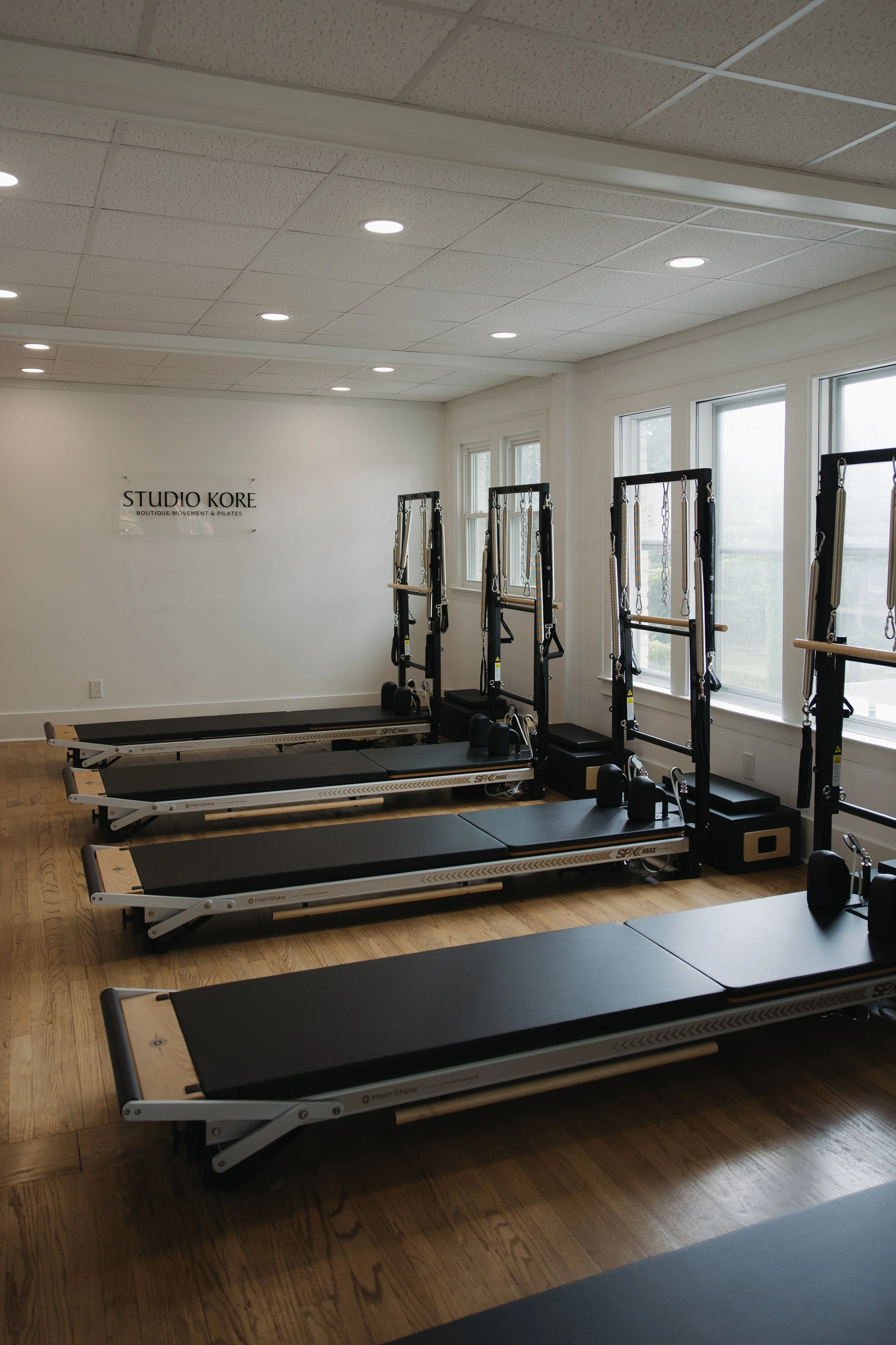 Pilates reformer machines lined up in a bright fitness studio with large windows, white walls, and a sign that reads "STUDIO KORE BOUTIQUE MOVEMENT & PILATES."