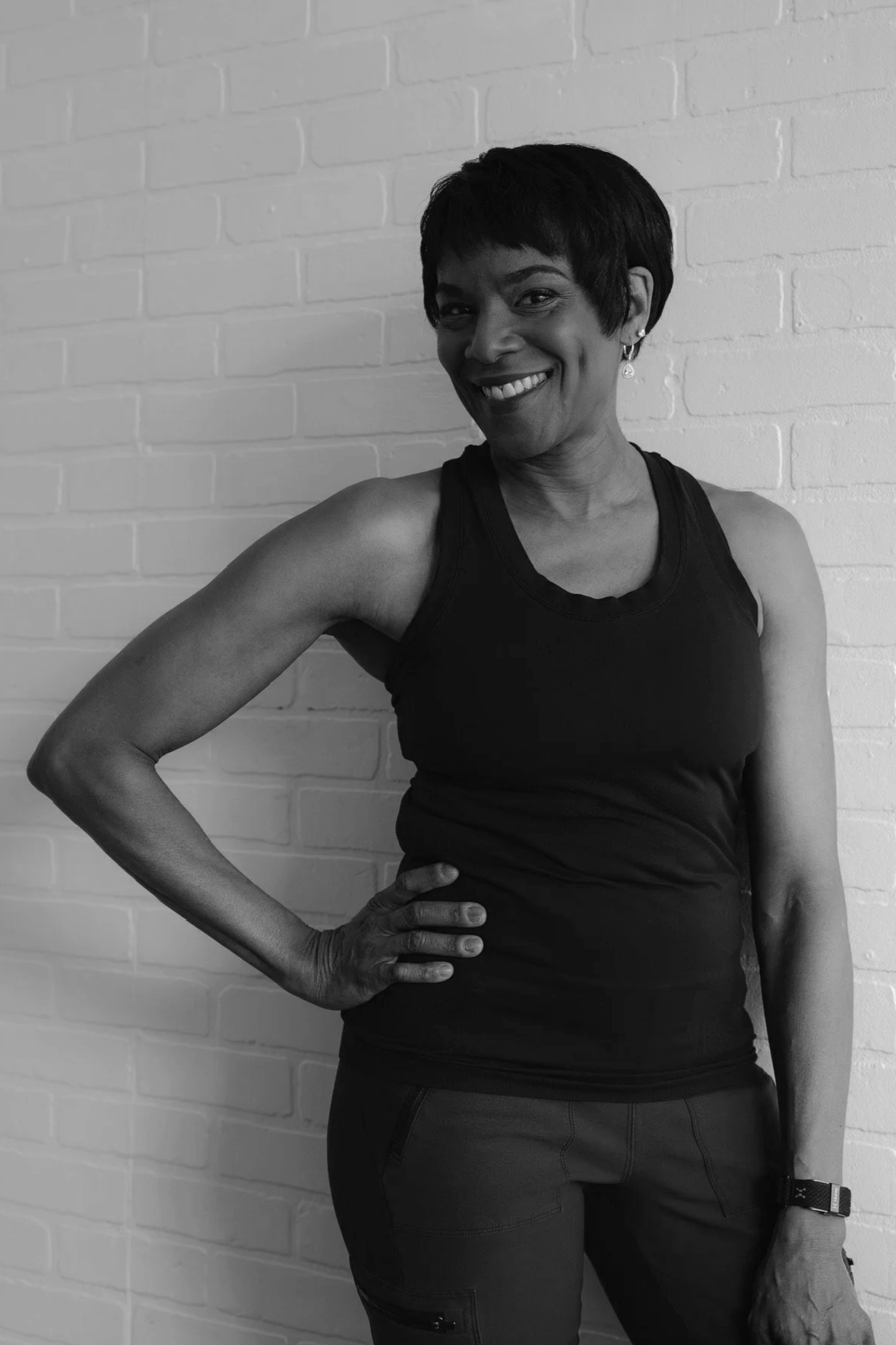 A woman with short hair smiling and posing with her hand on her hip against a brick wall, dressed in fitness attire.
