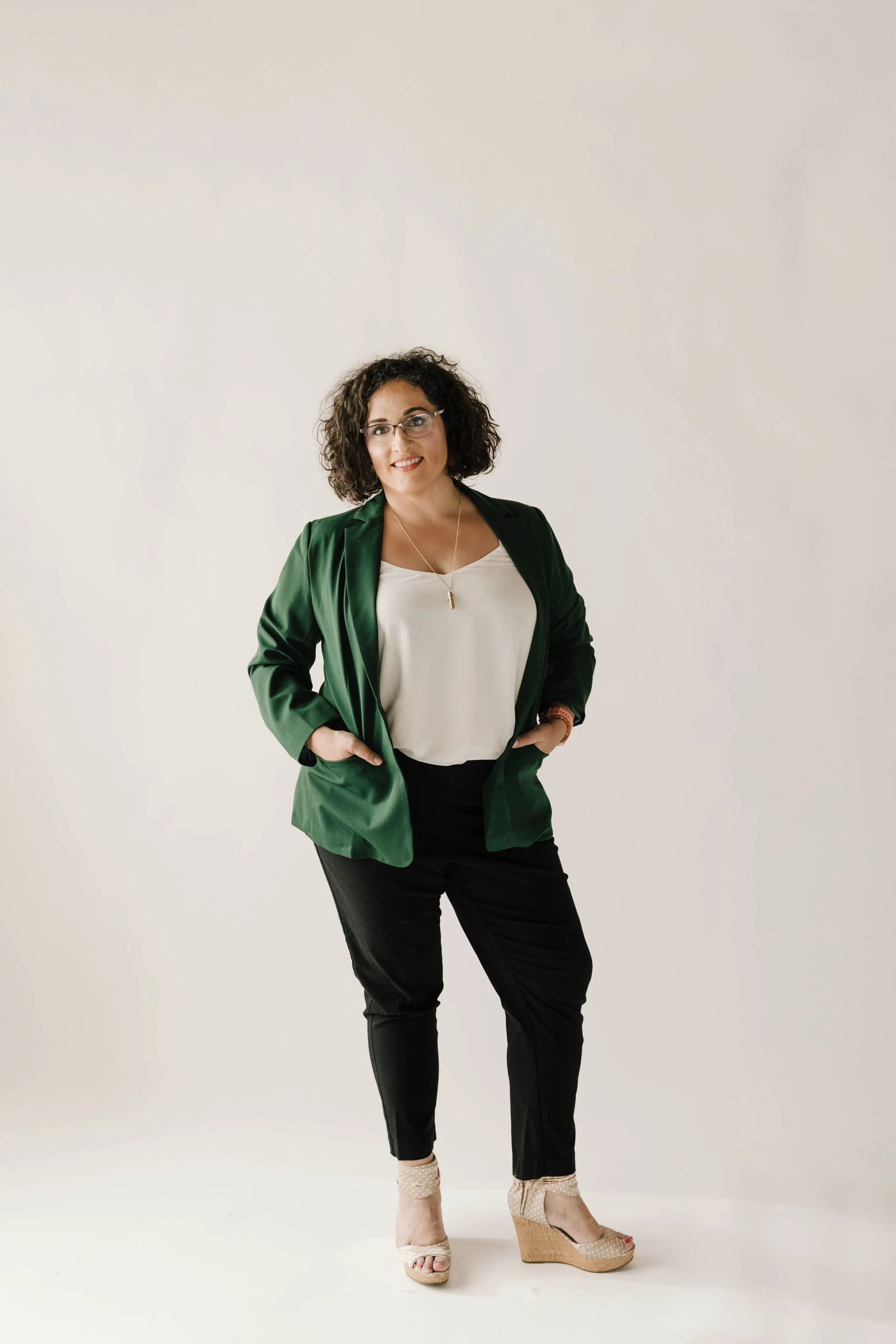 Woman with curly dark hair and glasses, wearing a green blazer, white blouse, black pants, beige wedge heels, smiling, standing against a plain white background.