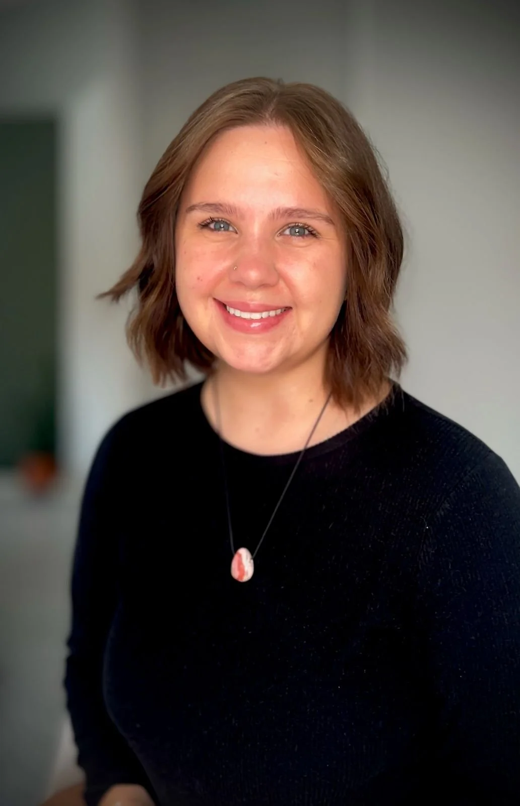 A woman with short brown hair, blue eyes, and a nose piercing smiling at the camera, wearing a black top and a necklace with a pink and white pendant.