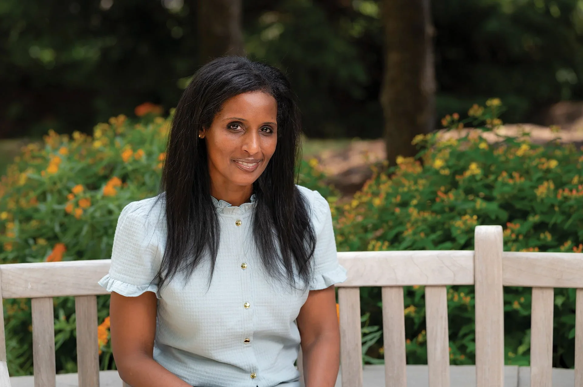 Dr. Birru Talabi sits on a park bench in front of two green bushes. Her long black hair sits on her shoulders as she wears a light blue top.