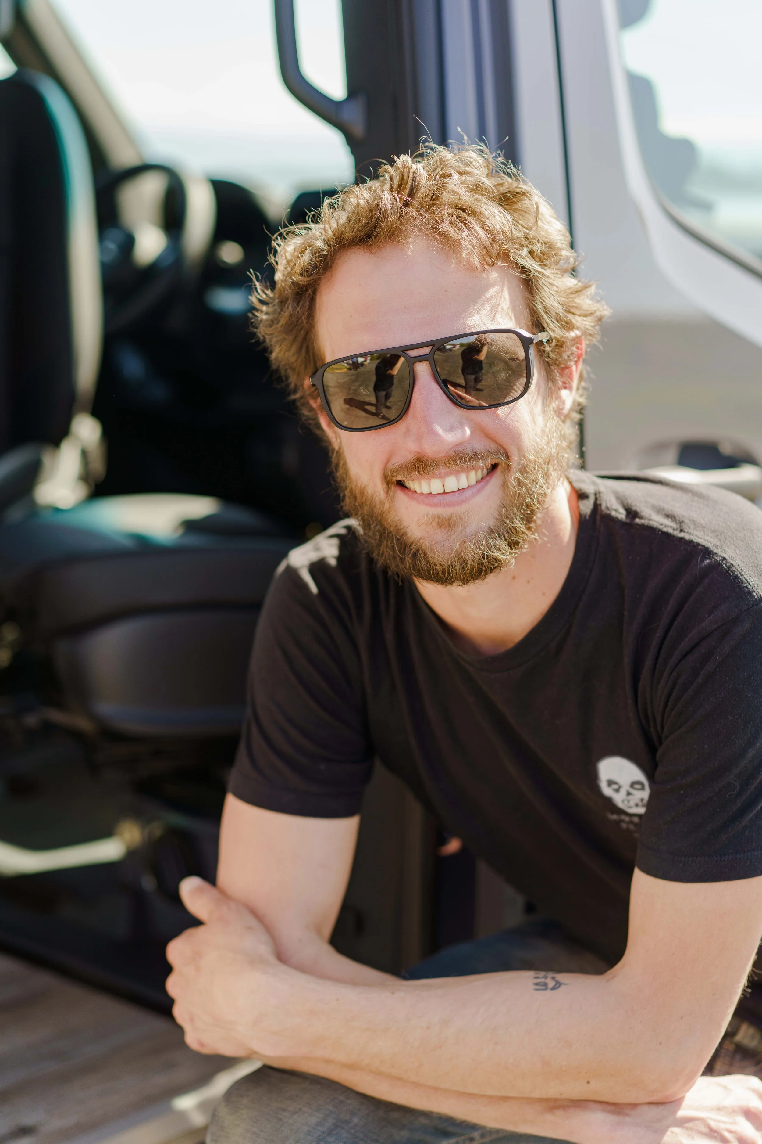 Man with sunglasses and beard sitting in open car door, smiling.