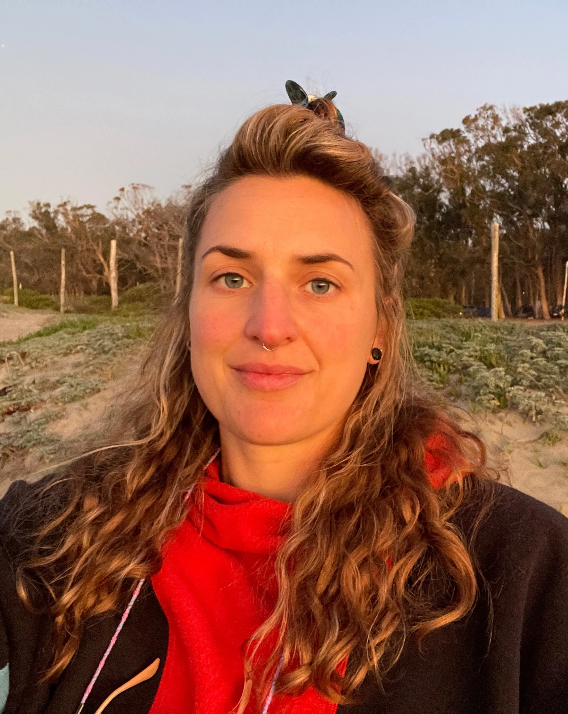 Person with curly hair wearing a red shirt and dark jacket, standing outside with trees and a sandy area in the background.