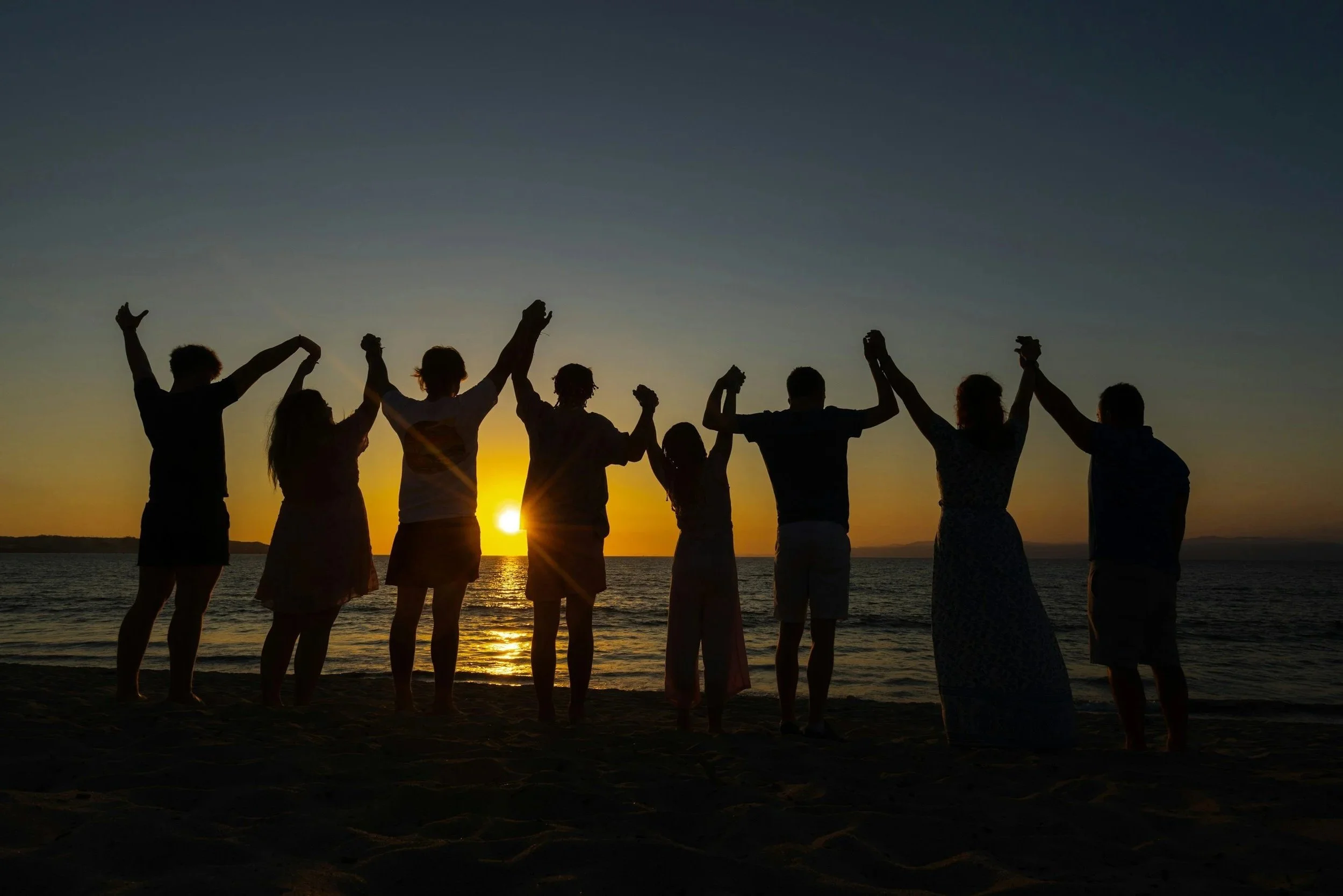 Group of people holding hands and raising them in celebration on a beach at sunset.