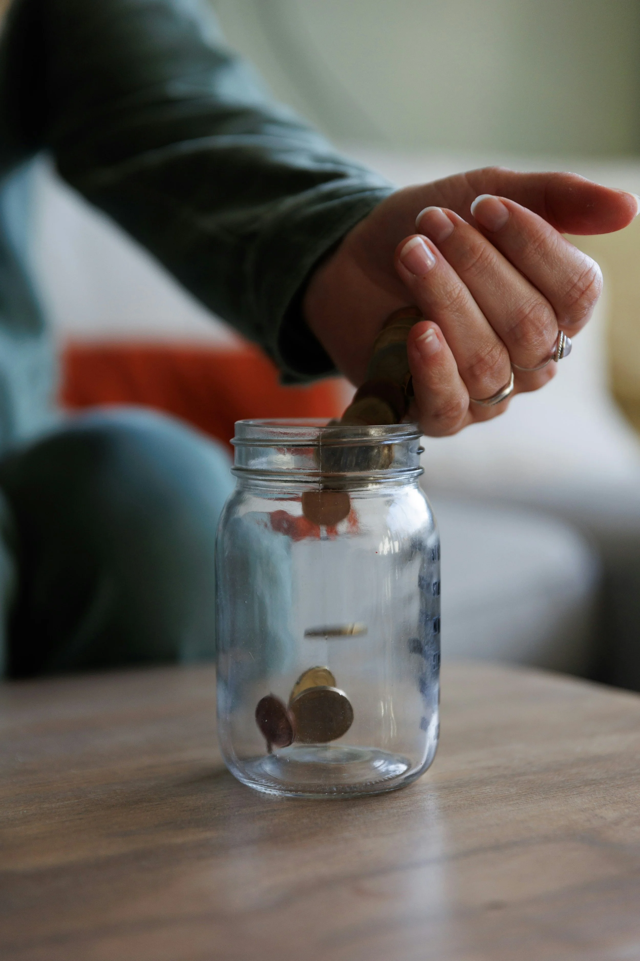 A person placing coins into a glass jar, which is sitting on a wooden surface.