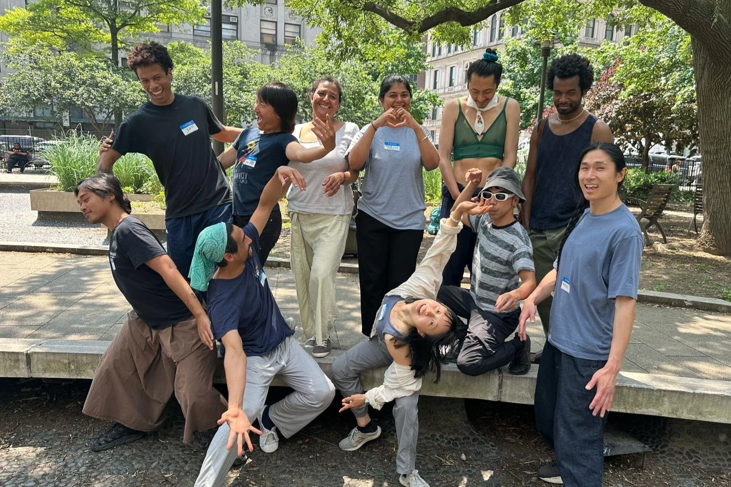 A diverse group of adults outdoors in a park, laughing and moving together in a playful, welcoming community practice.