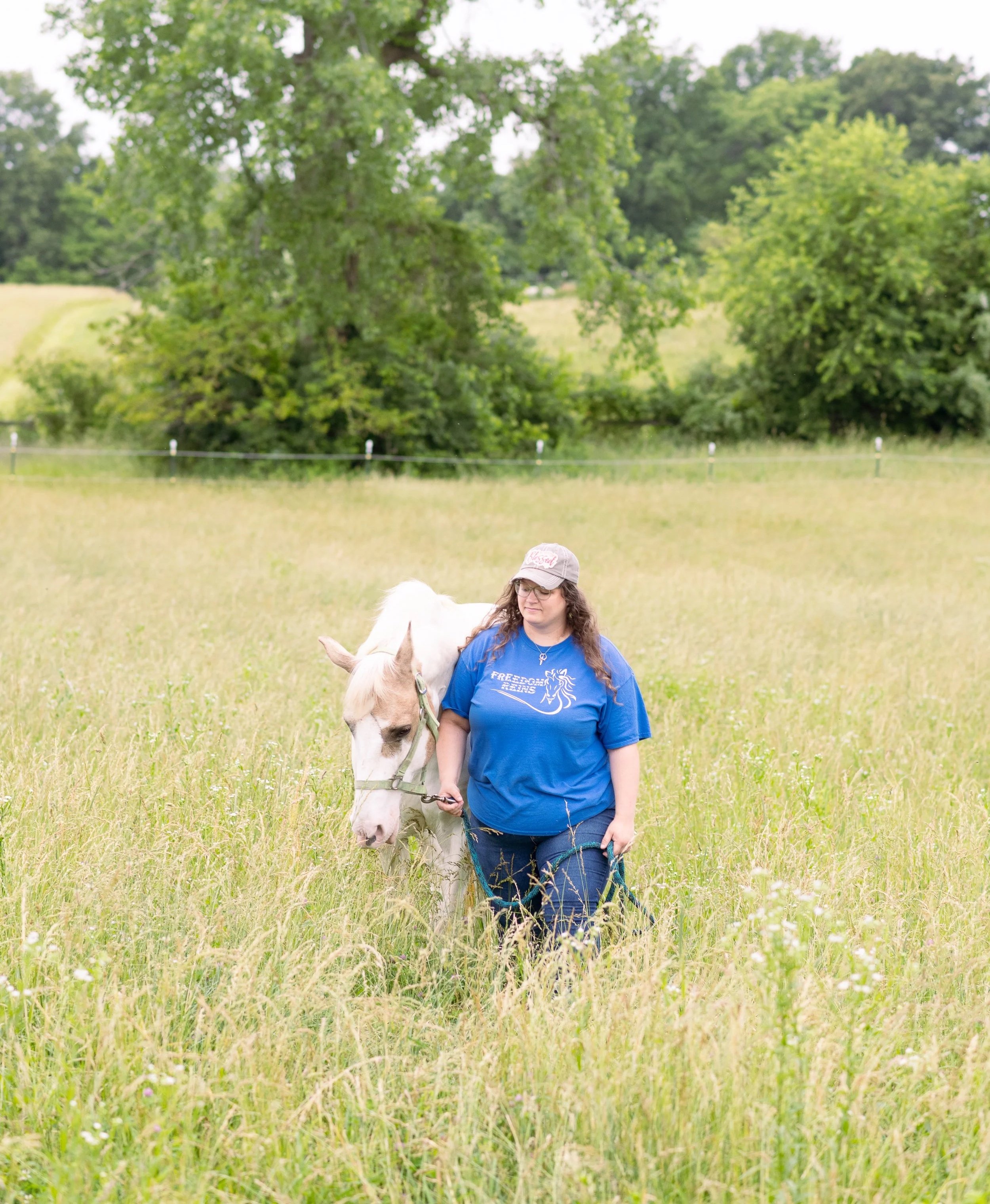 A woman with long curly hair, wearing glasses and a blue t-shirt, stands in a grassy field holding a leash attached to a white horse with brown patches. The woman is smiling, and there are tall trees and a fence in the background.
