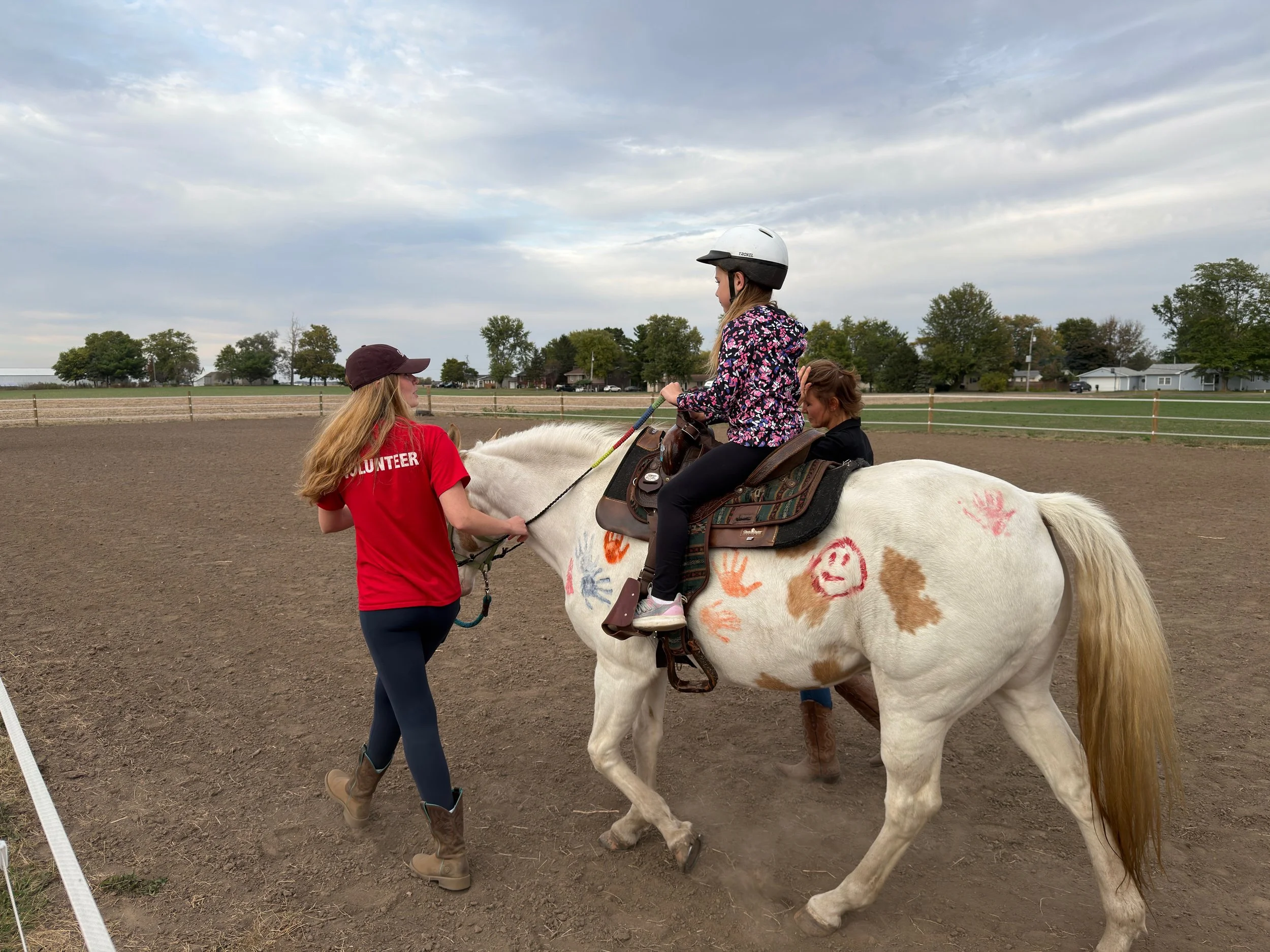 A young girl wearing a helmet and floral jacket riding a white horse decorated with handprints, accompanied by two women, one in a red volunteer shirt and another in a black jacket, in an outdoor riding arena.