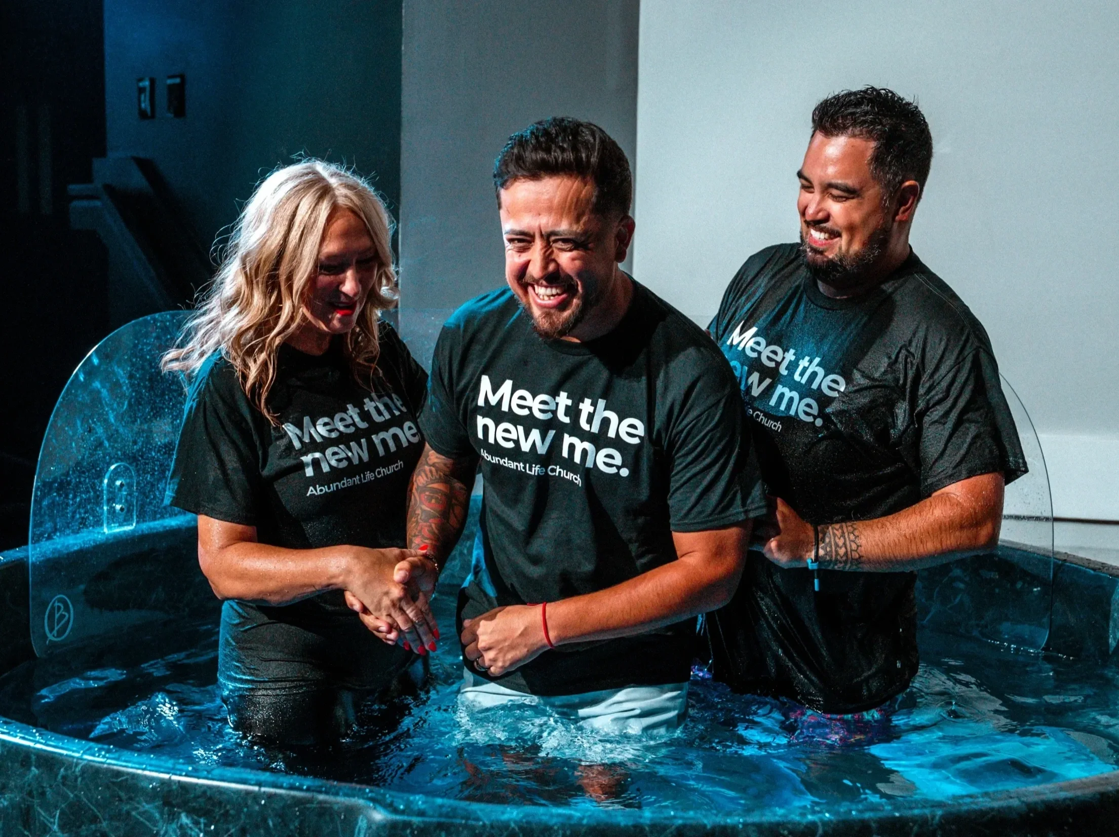 Three people standing in a baptism pool, smiling and celebrating. They are wearing black shirts with white text, and the woman in the center has wet hair and is touching her face with her hands.