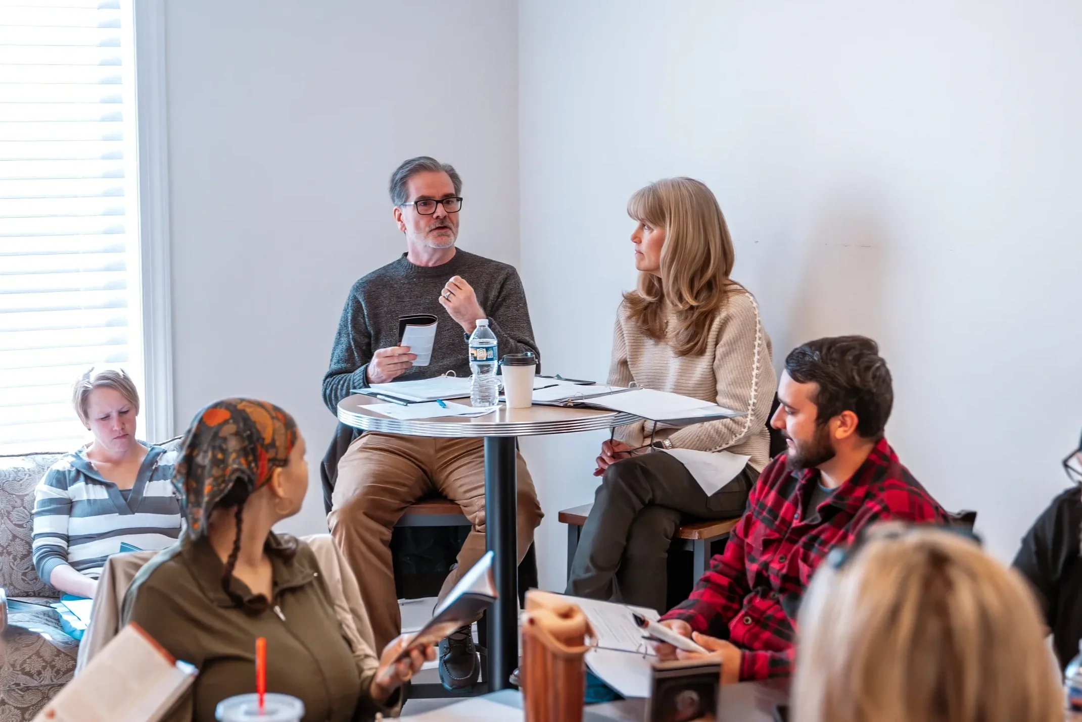People gathered around a table in a meeting room, engaged in discussion, with documents, notebooks, and digital devices on the table. A man in a blue shirt and a woman in a white shirt are visible, with others in the foreground and background, indicating an active group meeting or workshop.