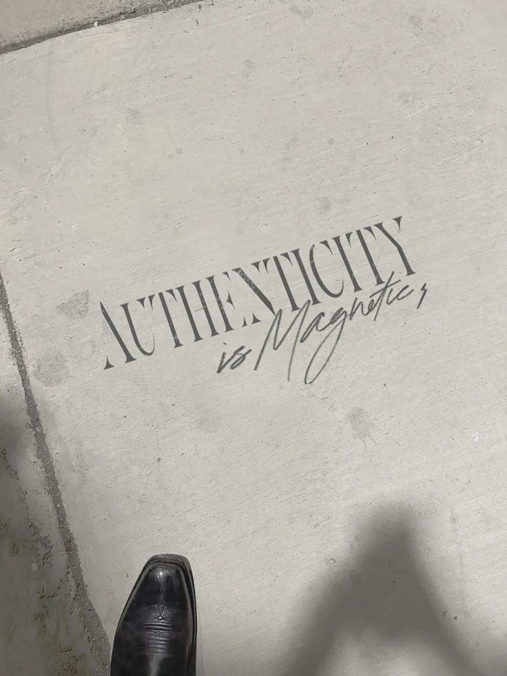 A concrete sidewalk or floor with a logo or sign that reads 'AUTHENTICITY is Magnetic' in stylized font. A person's black shoe is visible at the bottom of the image. Custom hand lettering and brand identity.