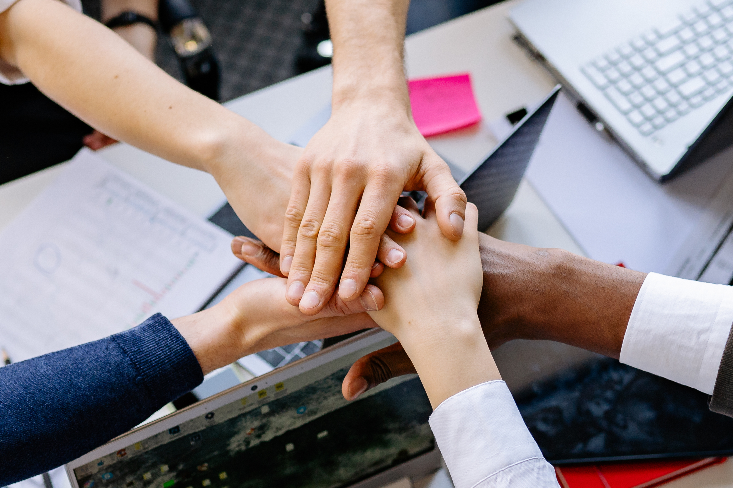 Five people placing their hands together in a team huddle over a desk with a laptop, documents, and sticky notes.