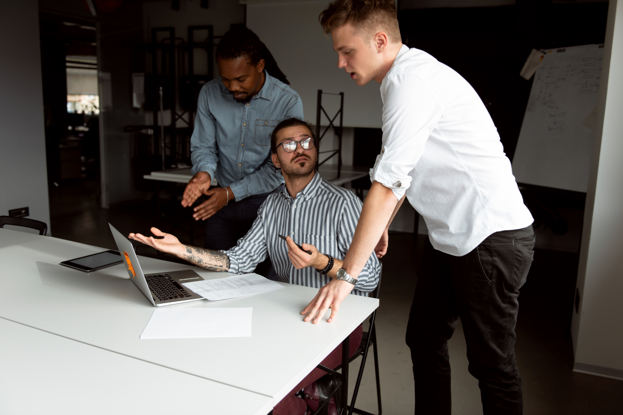 Three men having a discussion in a modern office. One seated at a table with a laptop, another standing and leaning toward him, and the third standing in the background.