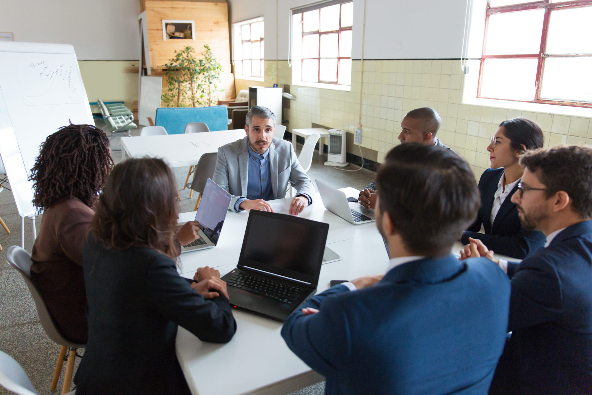 Business meeting in a bright conference room with seven people sitting around a white table, some using laptops, engaged in discussion.