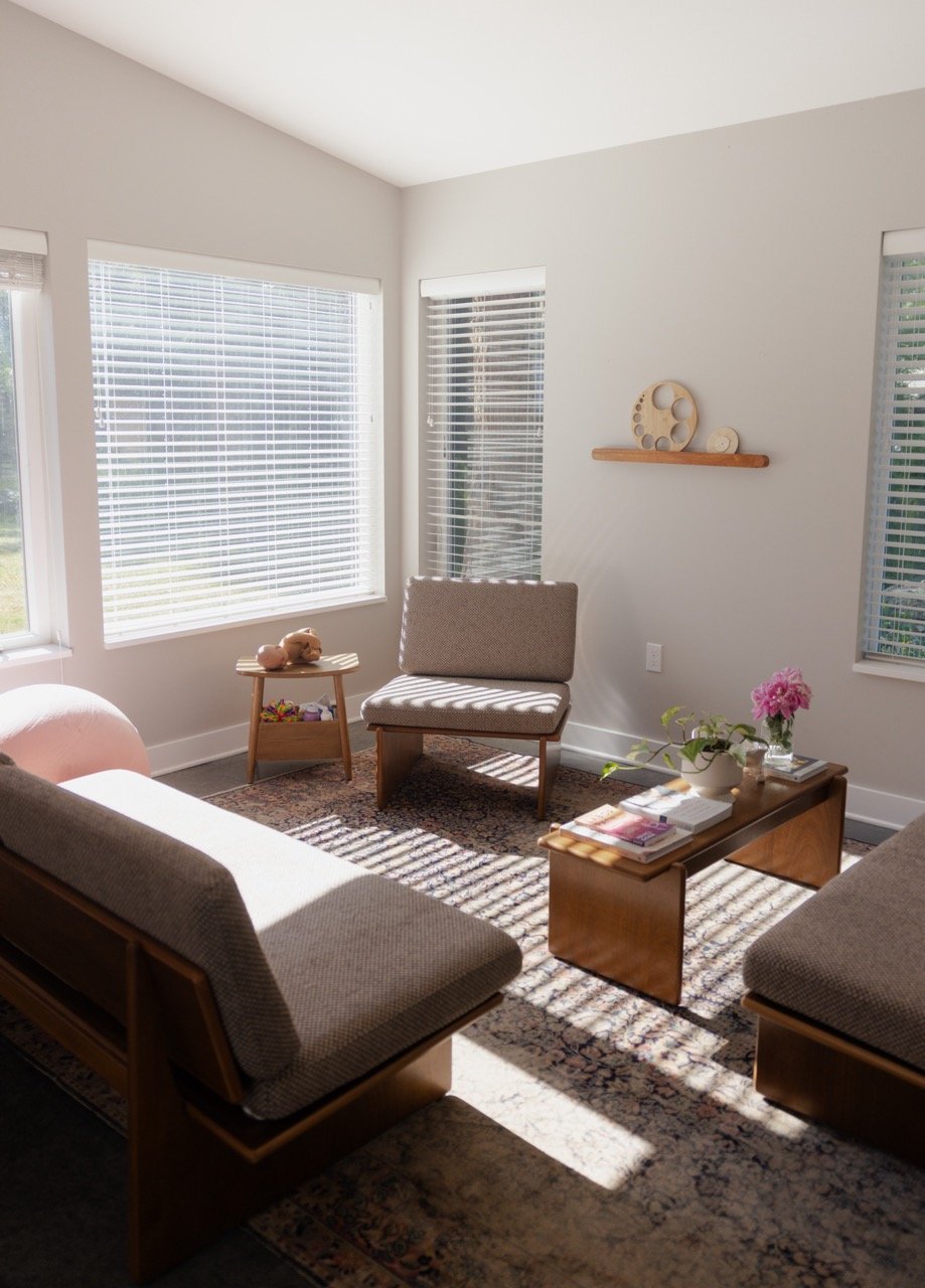 A cozy living room with large windows, a beige sofa, a chair, a wooden coffee table with magazines and a vase of pink flowers, a small side table with a toy, and decorative wall art, illuminated by natural sunlight.
