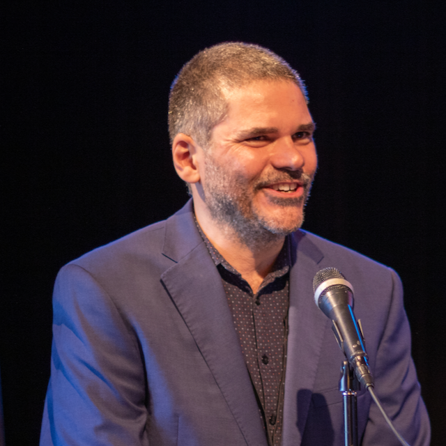 profile photo of Quinten Sheriff, who is wearing a button down shirt with a blue blazer. He has short brown hair and is speaking in front of a microphone.