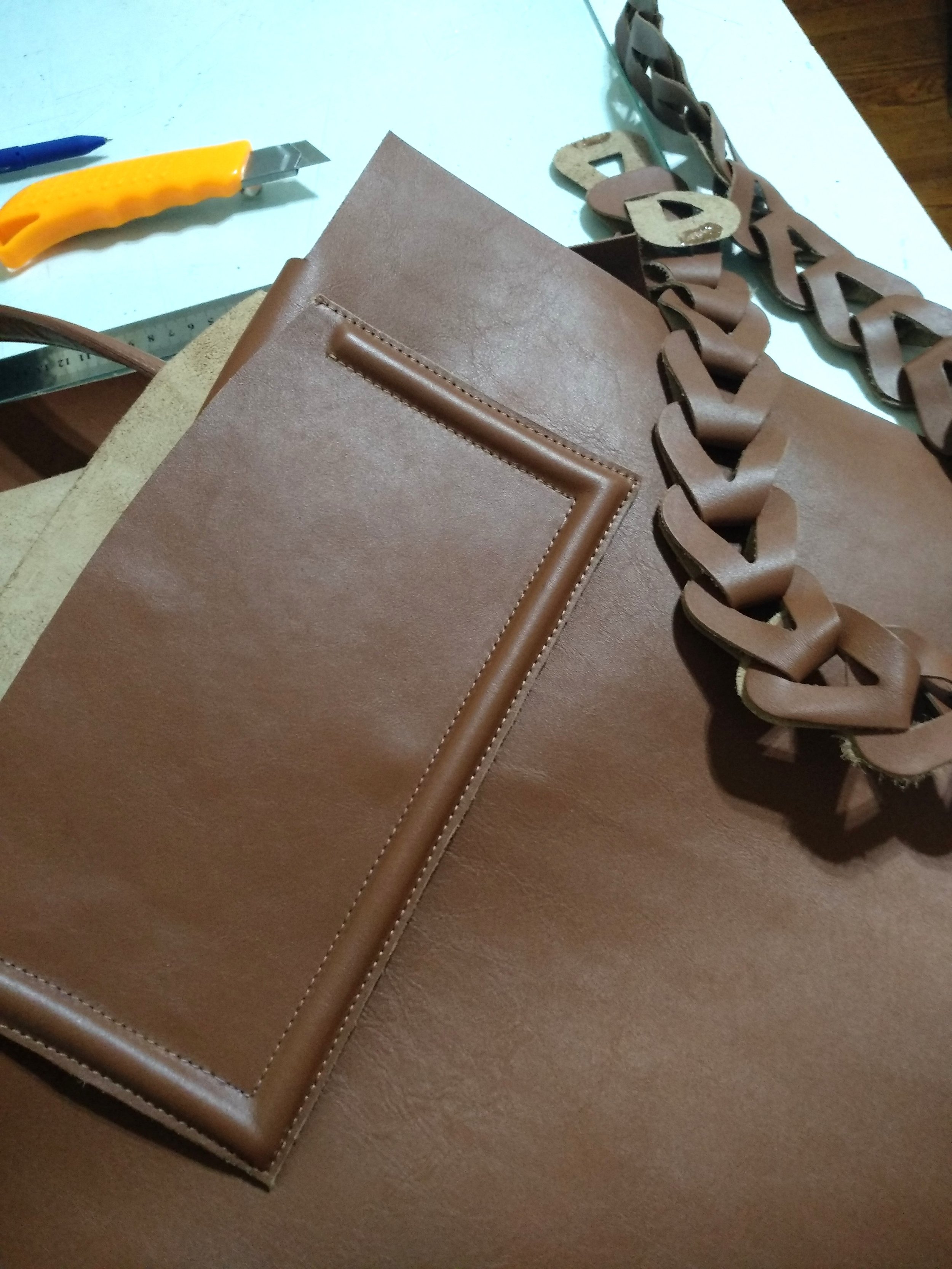 Brown leather materials and tools for crafting a handbag, including a chain, a pocket, and a utility knife on a work surface.