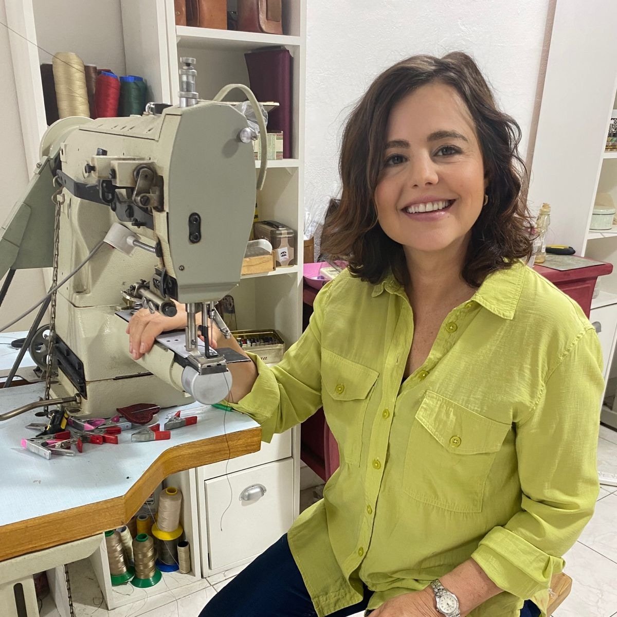 A woman leather artisan ssitting at a workbench with a sewing machine, smiling at the camera, in a craft room with shelves of thread and supplies.