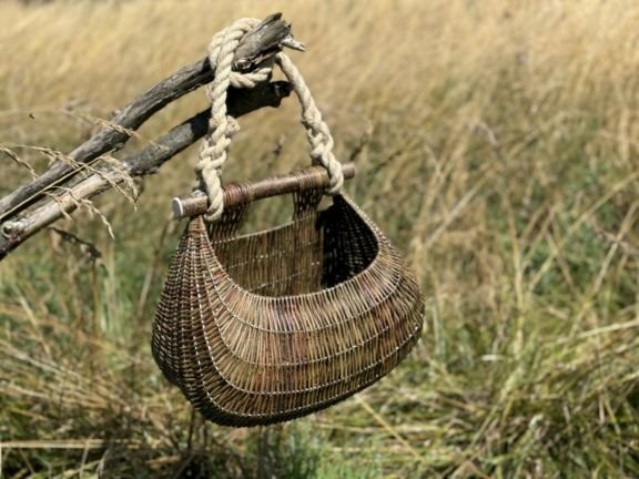 Handmade wicker basket with rope handle hanging on a branch, set against a sunlit field in the background.
