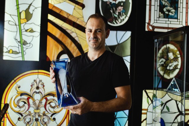A artisan holding a blue glass sculpture inside an art gallery with stained glass windows in the background.