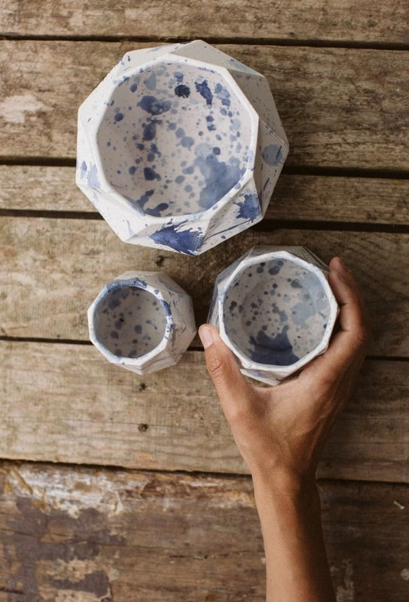 Three handmade ceramic cups with blue splatter paint resting on a wooden surface, one held in a hand.