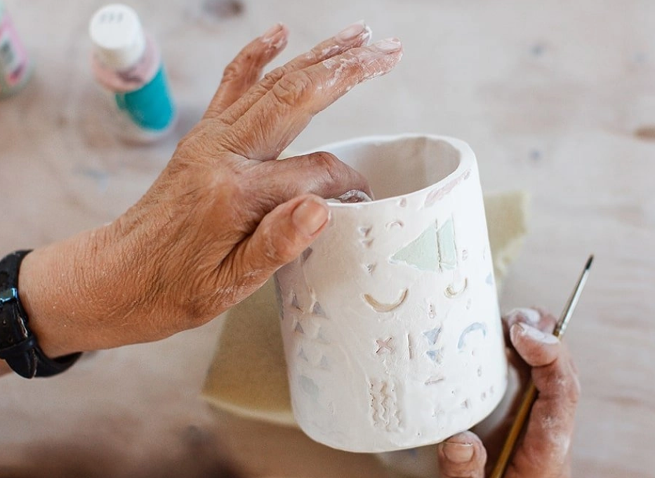 A person is hand-painting or decorating a white ceramic mug with carved or embossed patterns using a small paintbrush, on a wooden surface. In the background, there are bottles of paint.