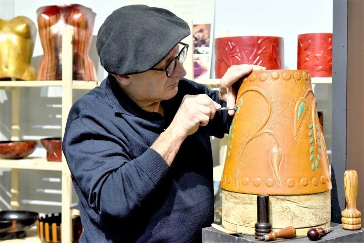 A leather craftsman wearing glasses and a dark shirt is carving and decorating a large, orange leather drum with turquoise accents, working carefully with a small tool.