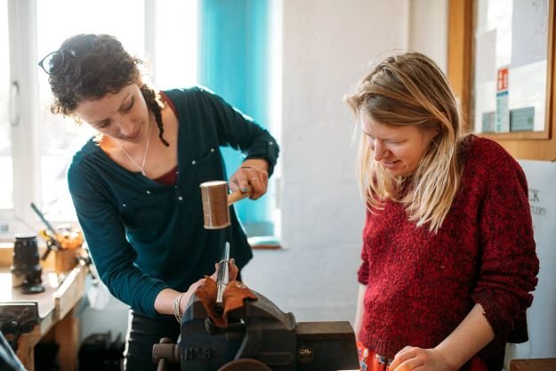 A woman is hammering a piece of wood while another woman watches, inside a workshop with tools and supplies around.