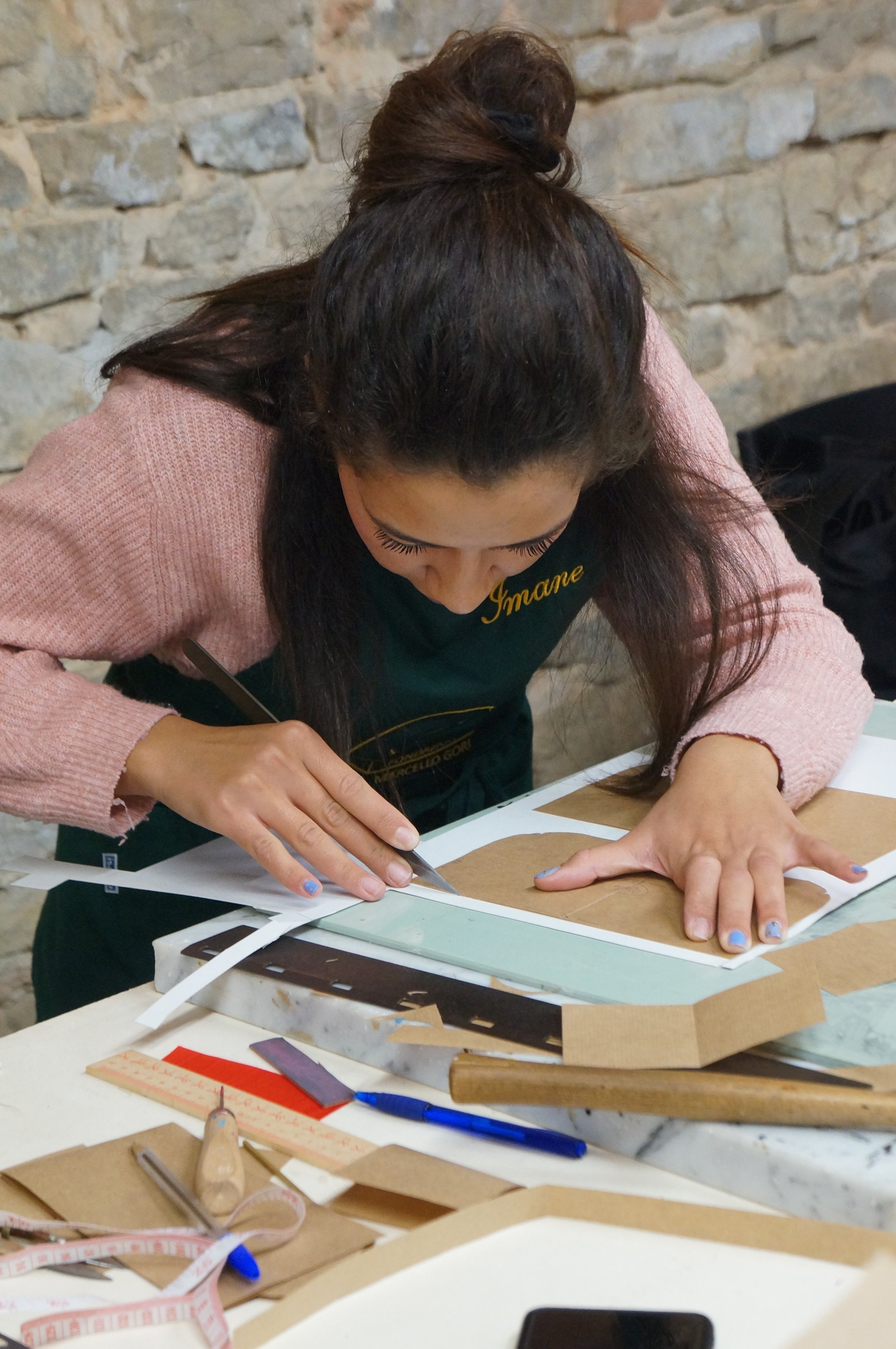 A woman with long dark hair tied in a bun, wearing a pink sweater and a dark apron, is leaning over a workspace and cutting leather with a craft knife. The workspace has tools, measuring tape, a ruler, and paper on it. The background is a stone wall.