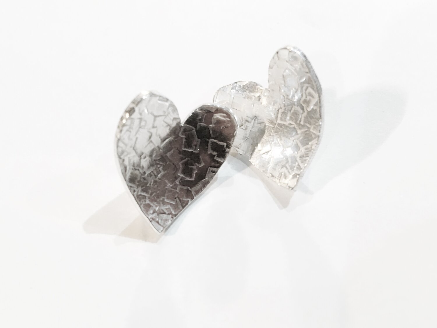 Handmade heart-shaped silver earrings with a textured, crackled surface, standing upright on a white background, casting soft shadows.