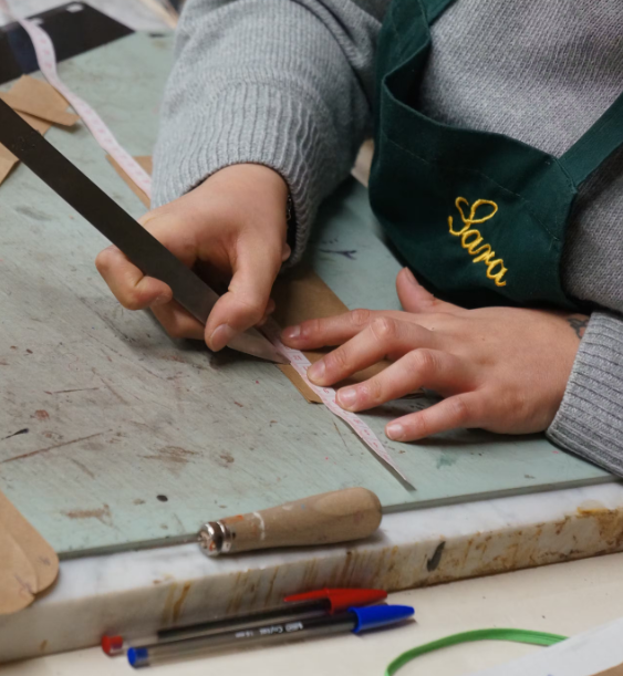 A person wearing a green apron with yellow embroidery working on a leather craft project, cutting a pink strip of leather with a straightedge cutter on a worktable.