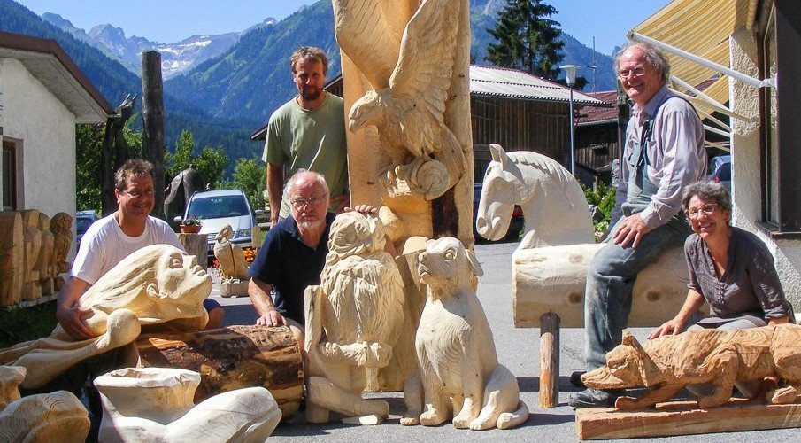 Group of six people outdoors with carved wood sculptures, mountains in the background.