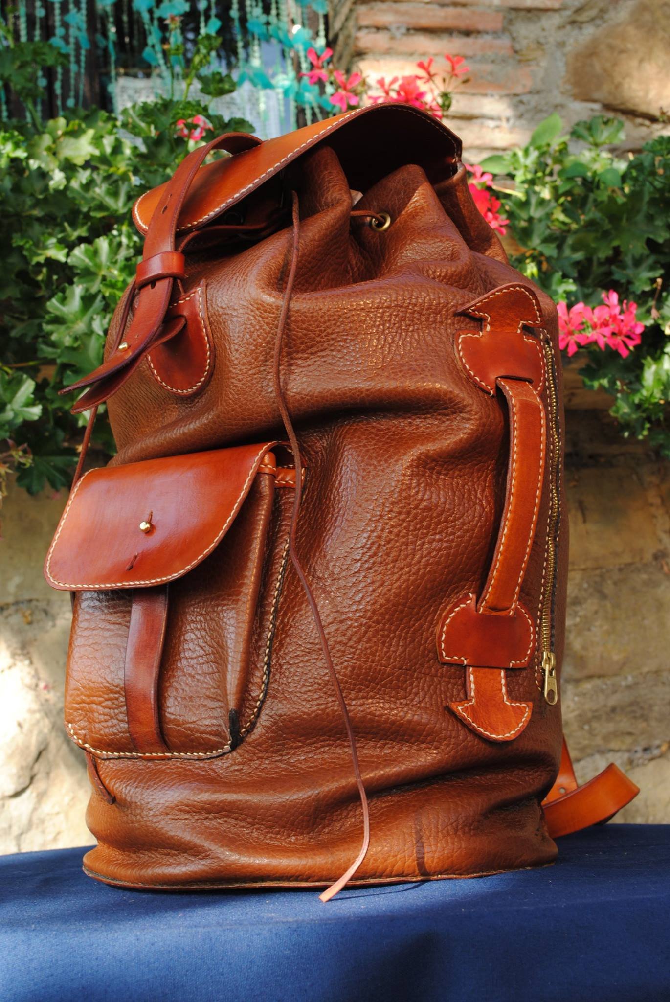 A large brown leather backpack with multiple pockets and a zipper, placed on a dark surface with a background of pink, green, and white flowers and greenery.
