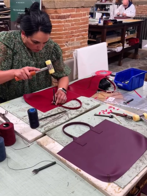 A woman working on a craft project at a table, using a hammer on a maroon bag with a brick wall in the background and another person working at a nearby station.