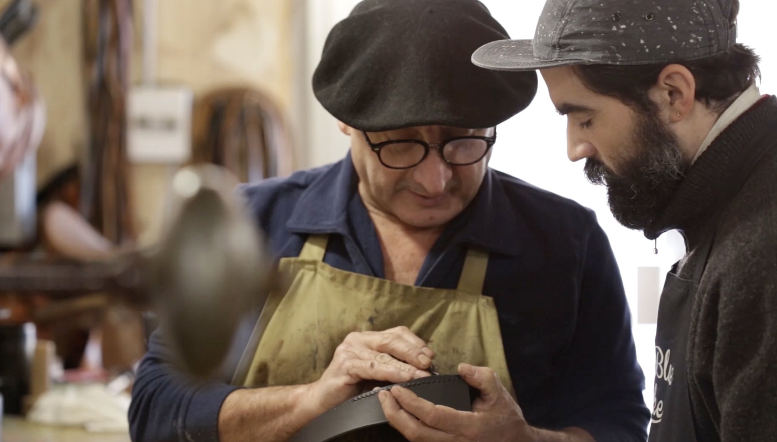 Two men are closely examining an object; one is wearing glasses, a beret, and an apron, and appears to be an artisan, while the other has a beard, a cap, and an apron.
