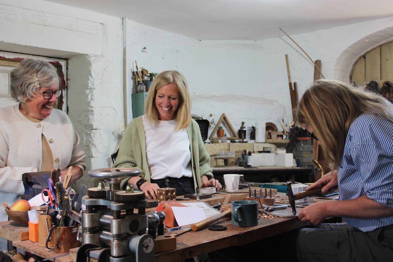 Three women are working and laughing in a jewelry making workshop with tools and materials on a wooden workbench.