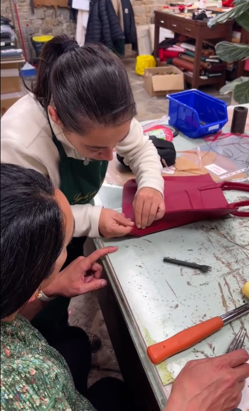 Two women working together on sewing a leather tote at a cluttered table in a workshop, with tools and leather nearby.