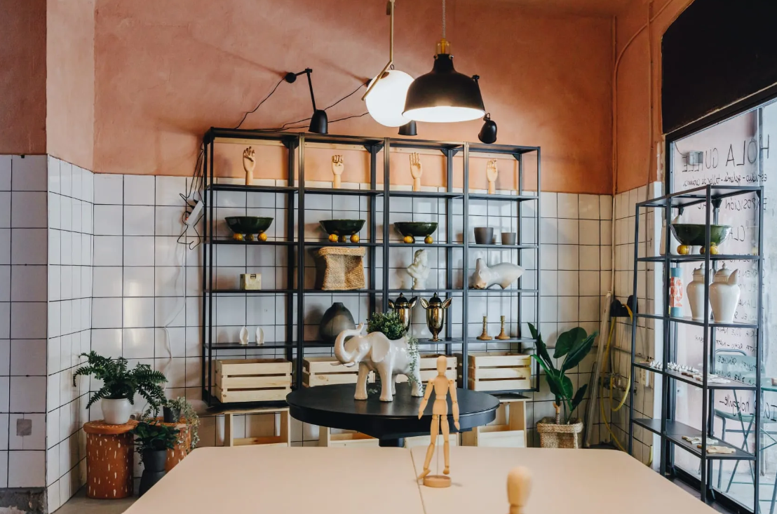 Interior of a stylish ceramic studio with shelves displaying handmade decorative objects and plants, and a round black table with animal and human figurines.