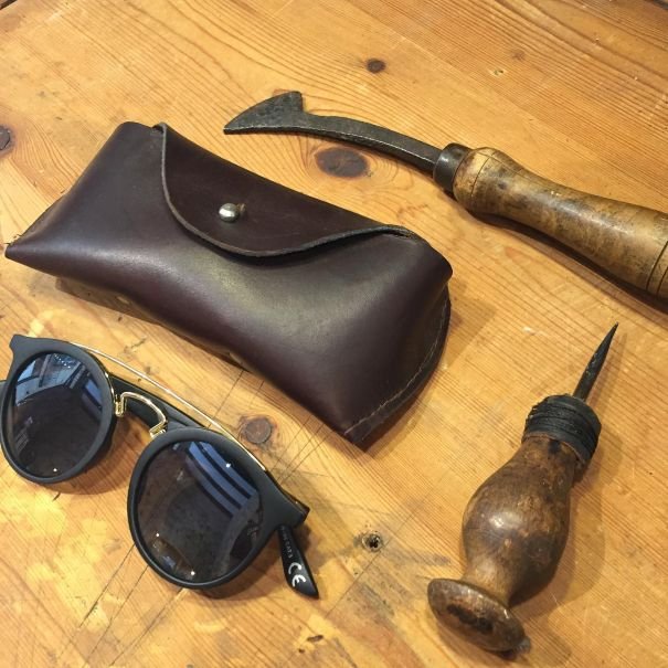Handmade leather glasses case on a wooden table, surrounded by leatherworking tools and a pair of sunglasses.