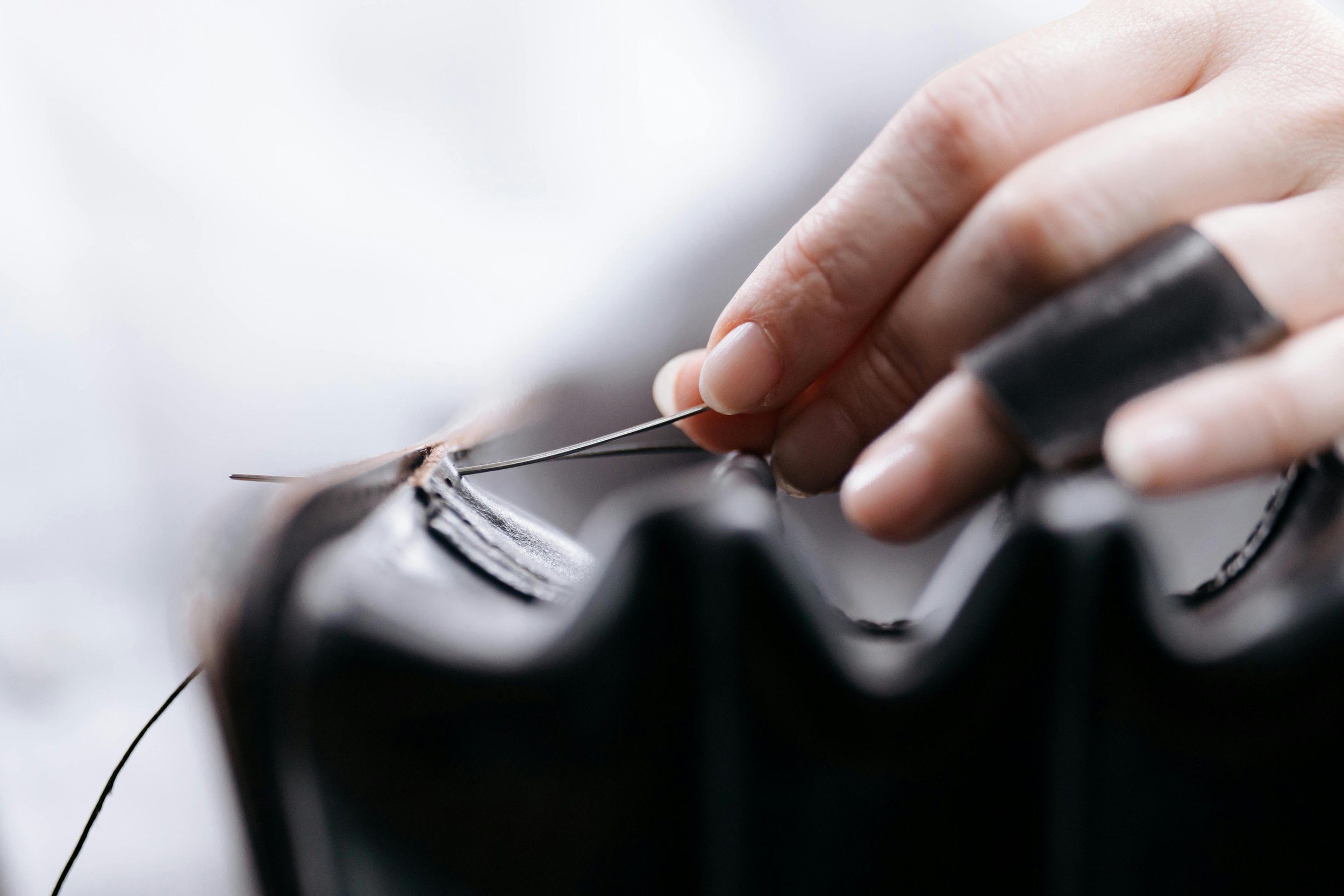 Close-up of a person sewing black leather material with a needle and black thread, using fingers protected by black tape.