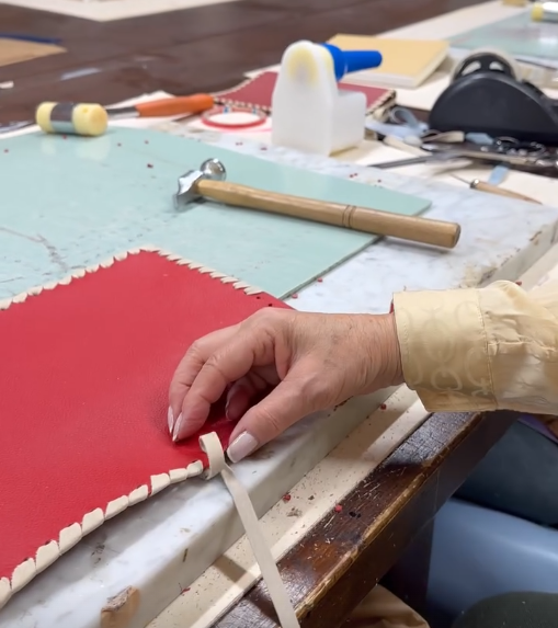 Close-up of a person's hand attaching a decorative edging to a red leather journal on a worktable, with tools and supplies scattered around.