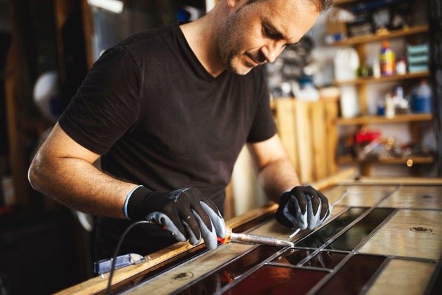 Man working on a stained glass project in a workshop, wearing black gloves and using a tool.