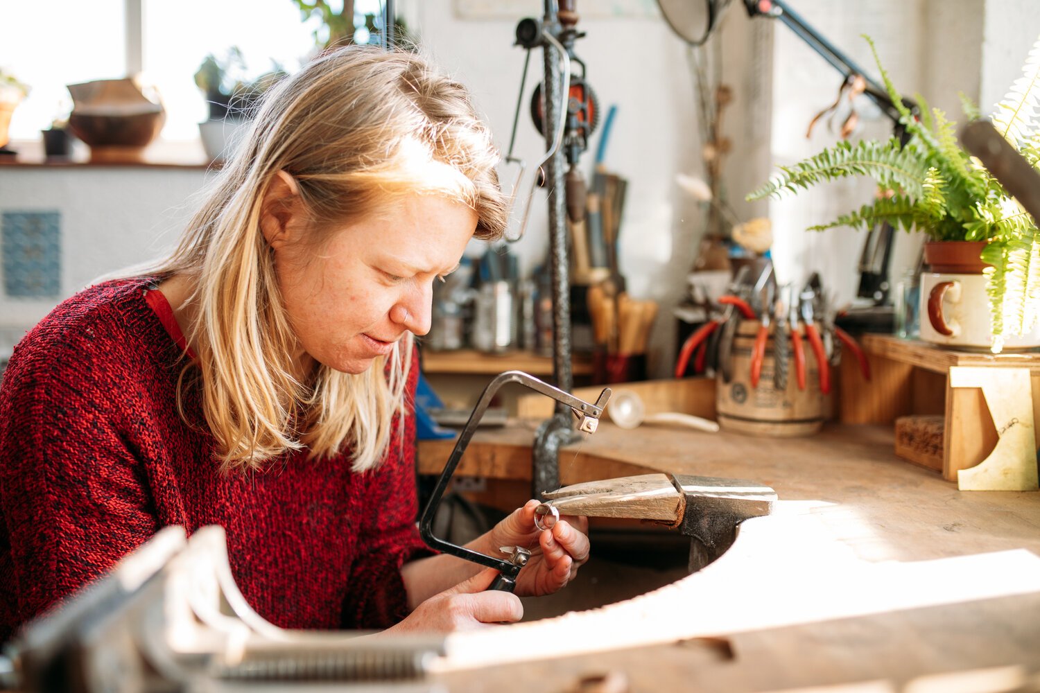 Woman in a red sweater working on a jewelry piece in her workshop with tools and plants around.